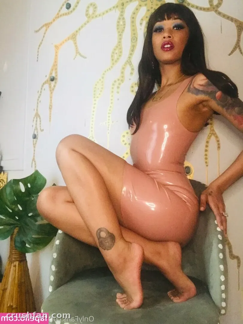 Skin Diamond con un vestido rosa brillante sentada en una silla de terciopelo verde contra una pared estampada