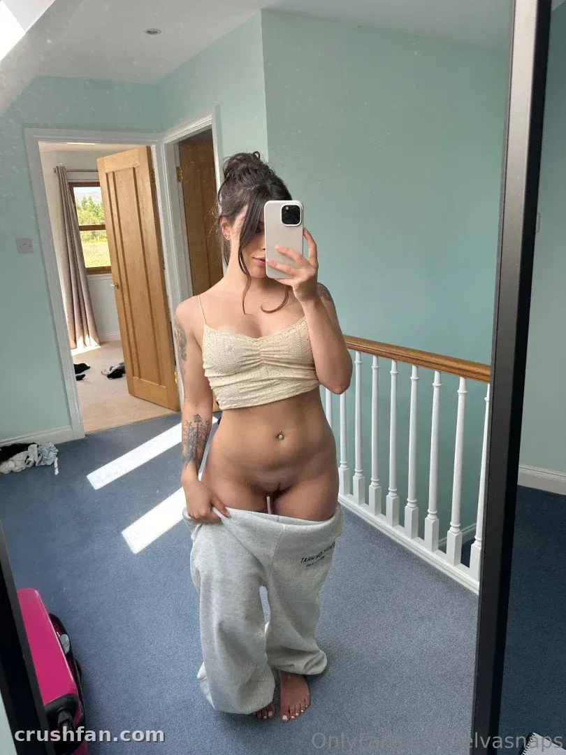 Elva Snaps tirando uma selfie no espelho em um corredor usando um top cropped e calça de moletom.