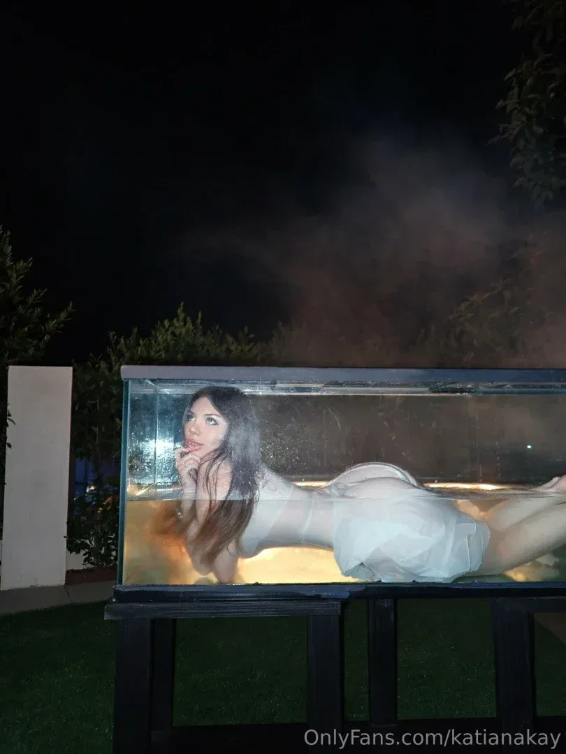 Katiana Kay reclining inside a glass tank outdoors at night
