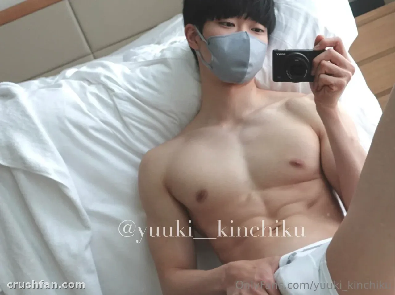 Yuuki lying on white bedding holding a small camera, wearing a grey face mask.