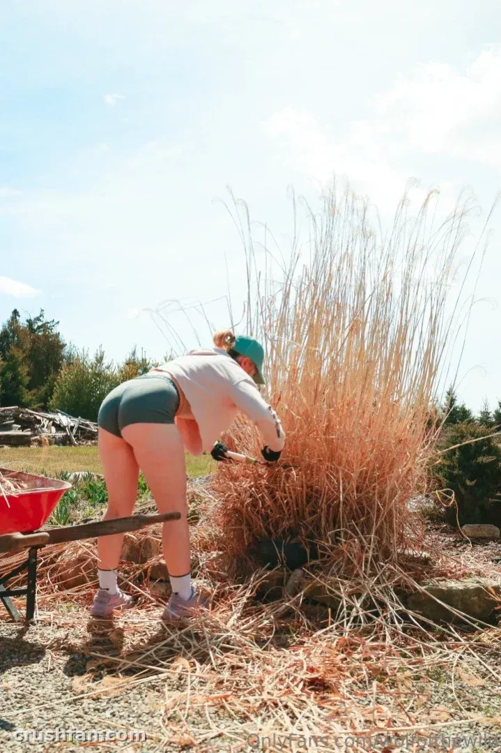 Holly Wolf in grüner Shorts und Kappe, die sich draußen neben einer roten Schubkarre über trockenes Gras beugt