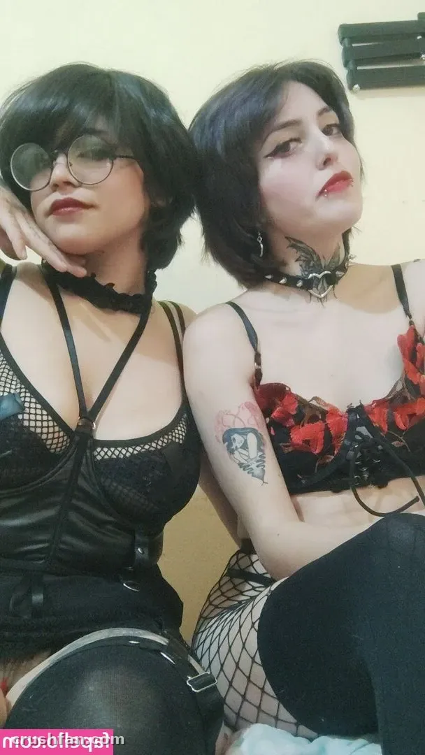 Deux femmes en tenues de dentelle sombre et chokers posent ensemble devant un mur clair.