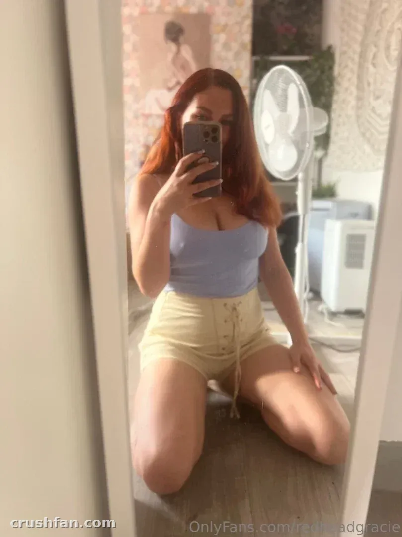 Grace Smyth kneeling on a wooden floor taking a mirror selfie in a light blue top and cream shorts.