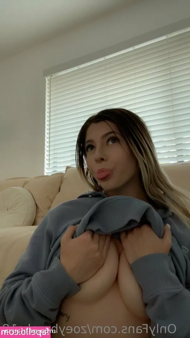 Zoey Brooks con una sudadera gris sentada en un sofá beige con persianas blancas detrás de ella