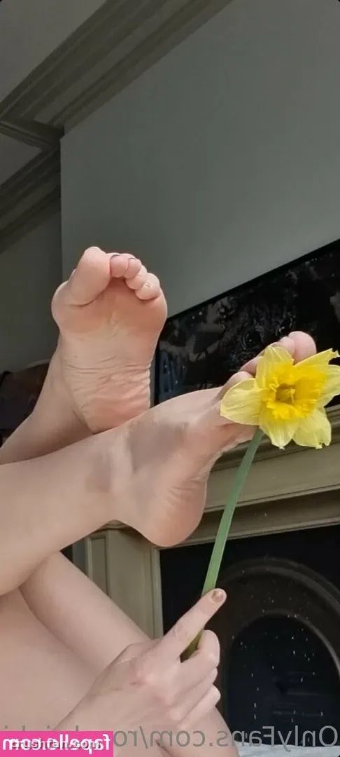 Rosa Brighid segurando um narciso amarelo ao lado dos pés descalços erguidos em ambiente interno