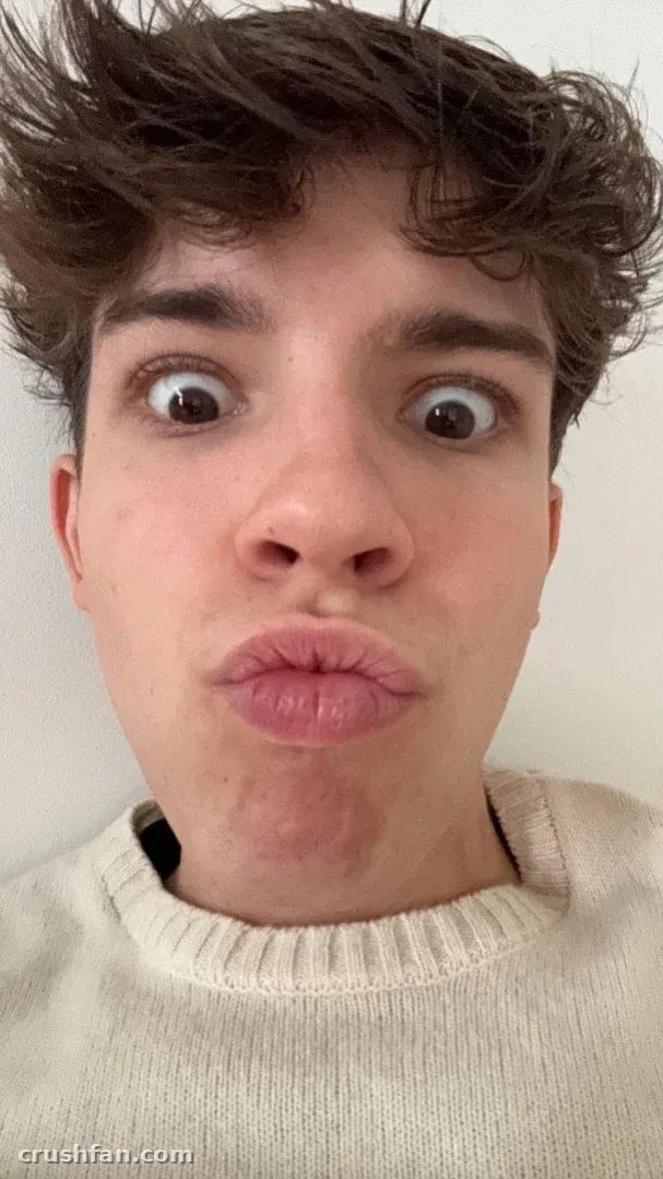 Selfie en gros plan de Manu dans un pull côtelé crème faisant une grimace espiègle avec de grands yeux contre un mur uni
