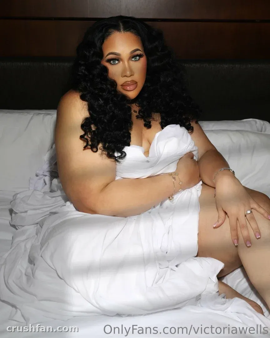 Victoria Wells con un vestido blanco sentada sobre ropa de cama blanca en interiores