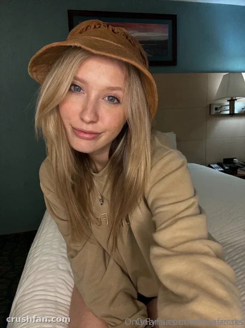 Ava dans un bob beige et un sweat à capuche assise sur un lit dans une pièce à l'éclairage doux
