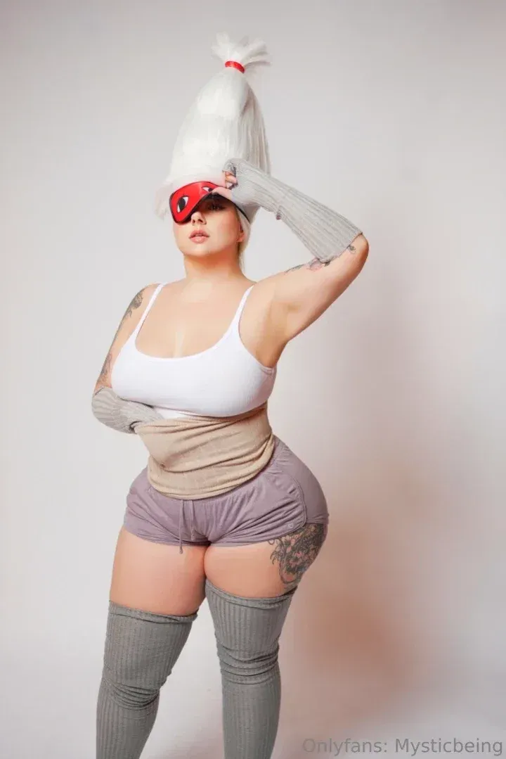 MysticBeing in einem weißen Top, roter Augenmaske und grauen Overknee-Socken vor einem schlichten Hintergrund