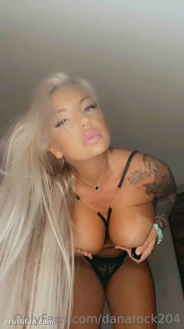 Dana Rock de lingerie preta e longos cabelos platinados contra uma parede cinza lisa