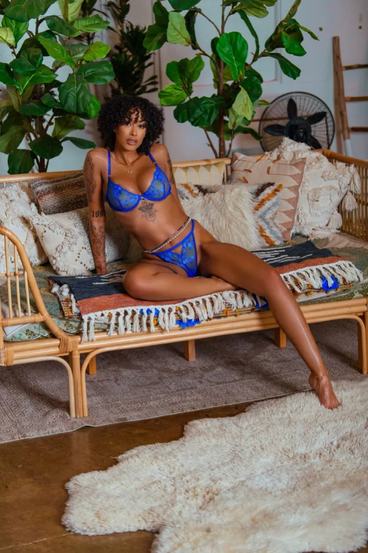 Miss B. Nasty in blue lace lingerie seated on a rattan daybed surrounded by plants.