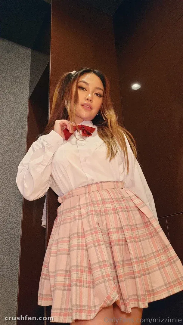Mizzi Mie in a white blouse and pink plaid skirt standing indoors