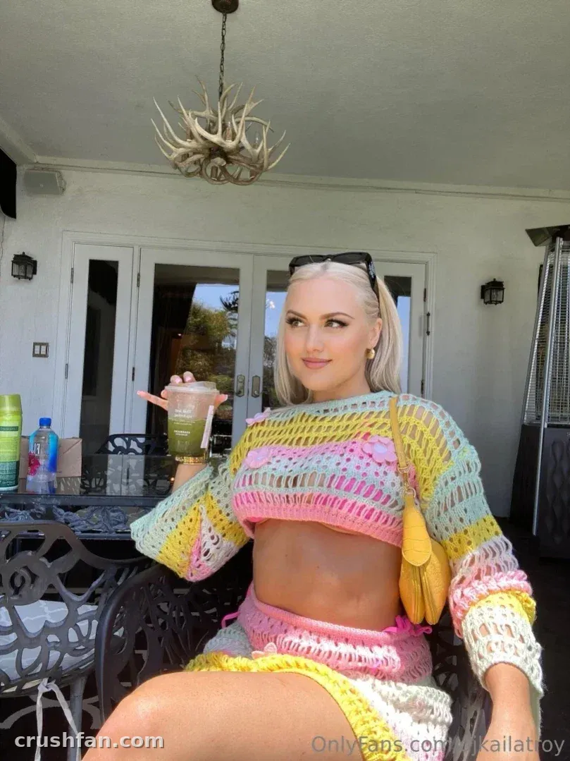 Kaila Troy sentada em um pátio com um top e saia de crochê coloridos, segurando uma bebida.