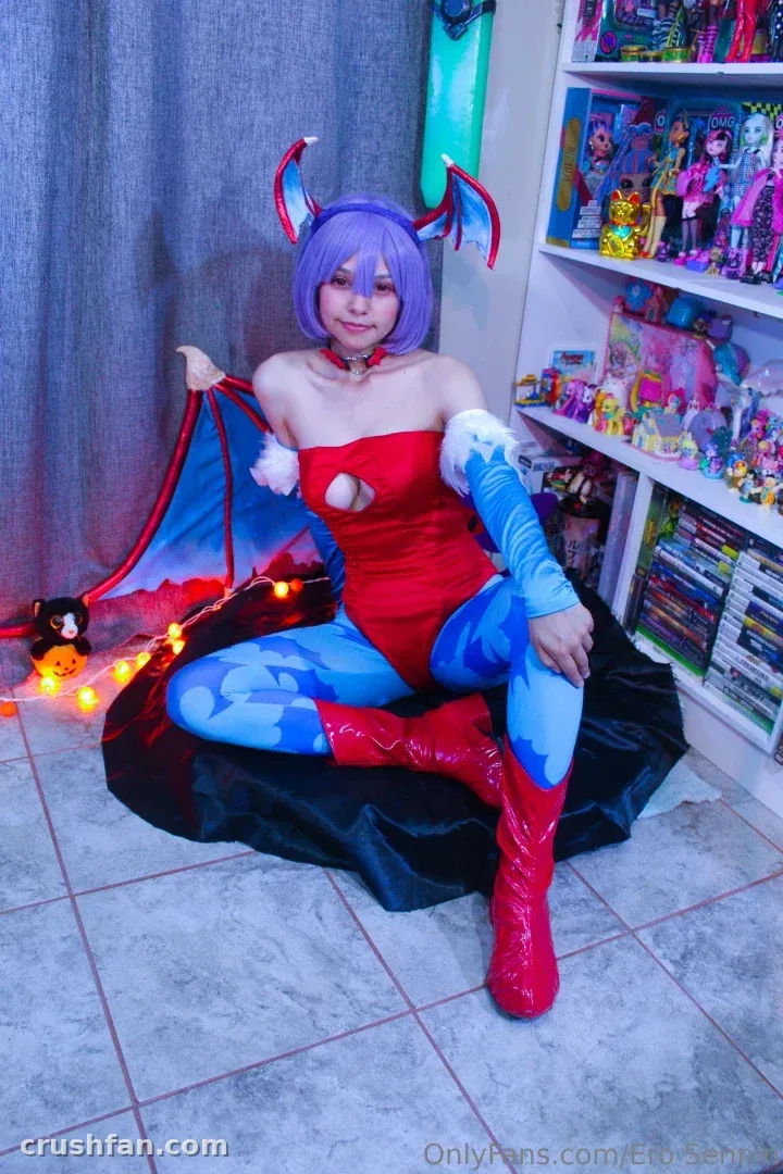 Cosplayeuse en bodysuit rouge et jambières bleues assise sur une surface sombre et drapée en intérieur.