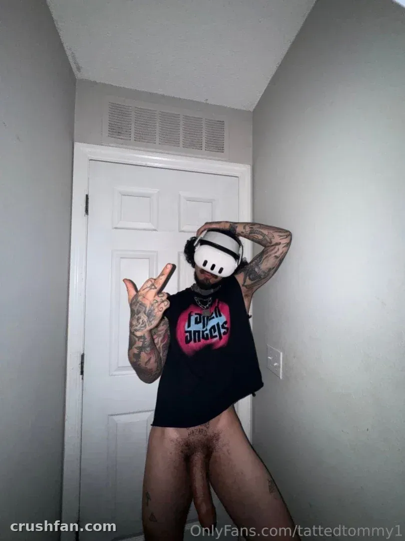 Tommy com uma regata gráfica preta e headset de realidade virtual posando em ambiente interno perto de uma porta branca.