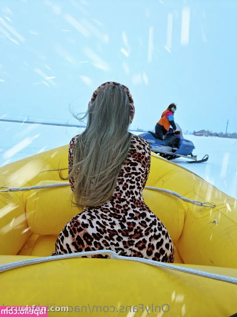 Criadora de maiô com estampa de leopardo sentada em uma boia amarela em um lago congelado com um snowmobile ao fundo.