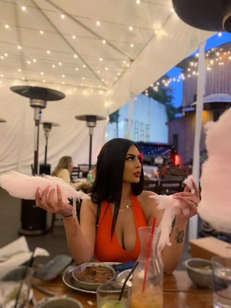 Woman in an orange top holding pink cotton candy at an outdoor table under string lights.