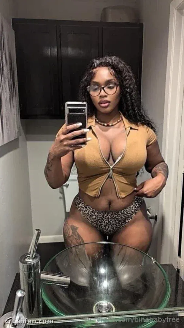 Sabrina Lynn tirando uma selfie no espelho em um banheiro usando um top mostarda e calcinha de biquíni de leopardo.