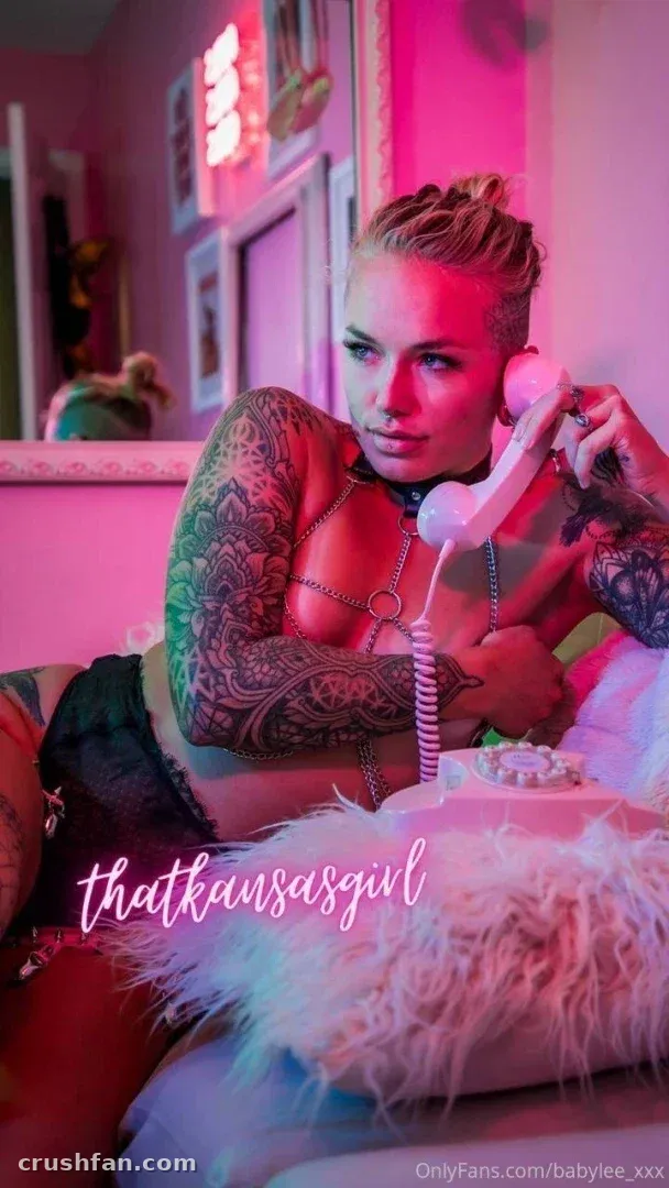 Créatrice en lingerie sombre tenant un téléphone vintage rose dans une pièce éclairée au néon avec des tatouages visibles.