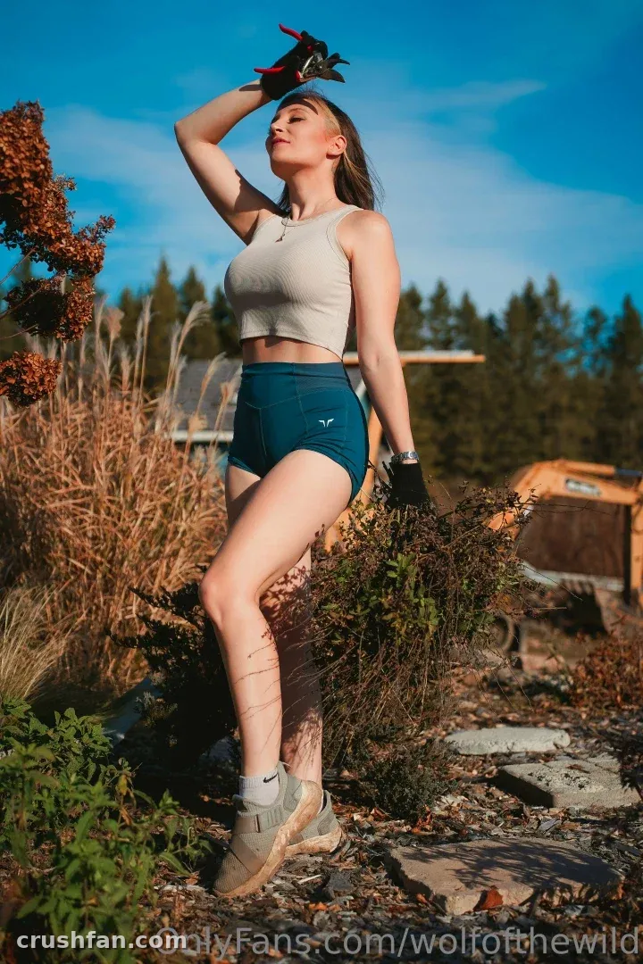 Holly Wolf in a beige crop top and teal shorts posing outdoors under a blue sky.