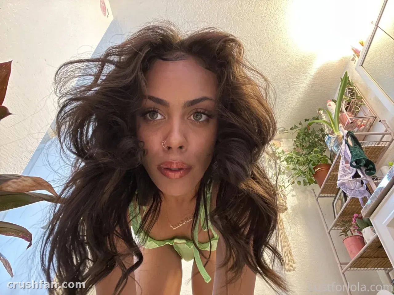Criadora de longos cabelos ondulados com blusa verde posando em ambiente interno perto de plantas em vasos