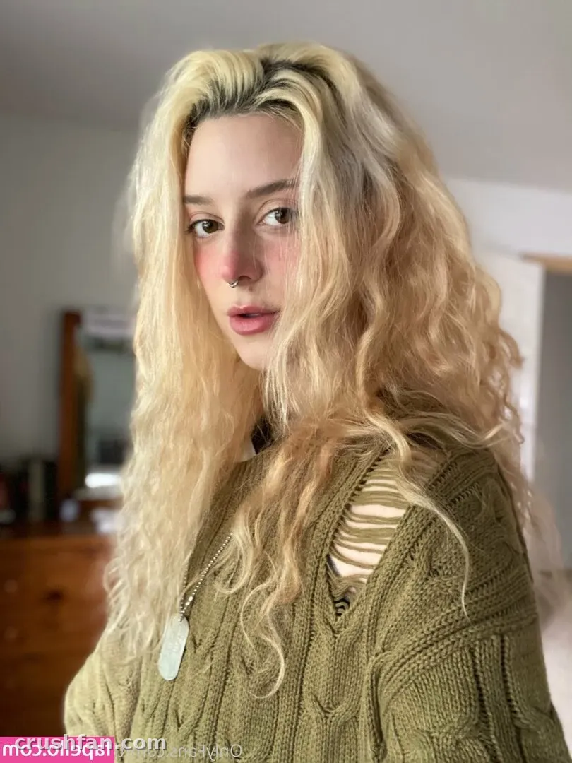 Cam Damage with long wavy blonde hair in a green knit sweater in a softly lit room.