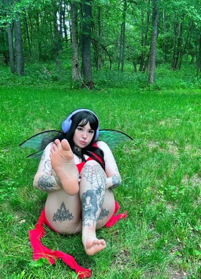 Venomous Dolly sits barefoot in a forest wearing fairy wings and headphones.