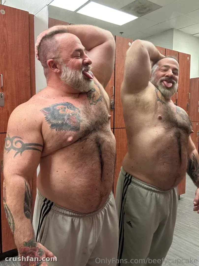 Shirtless bearded man with chest tattoos posing in a locker room mirror