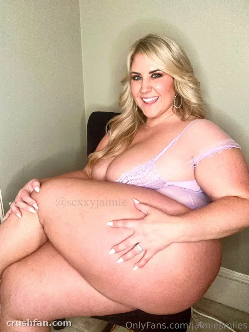 Juicy J seated indoors in lilac lace lingerie, smiling toward the camera.