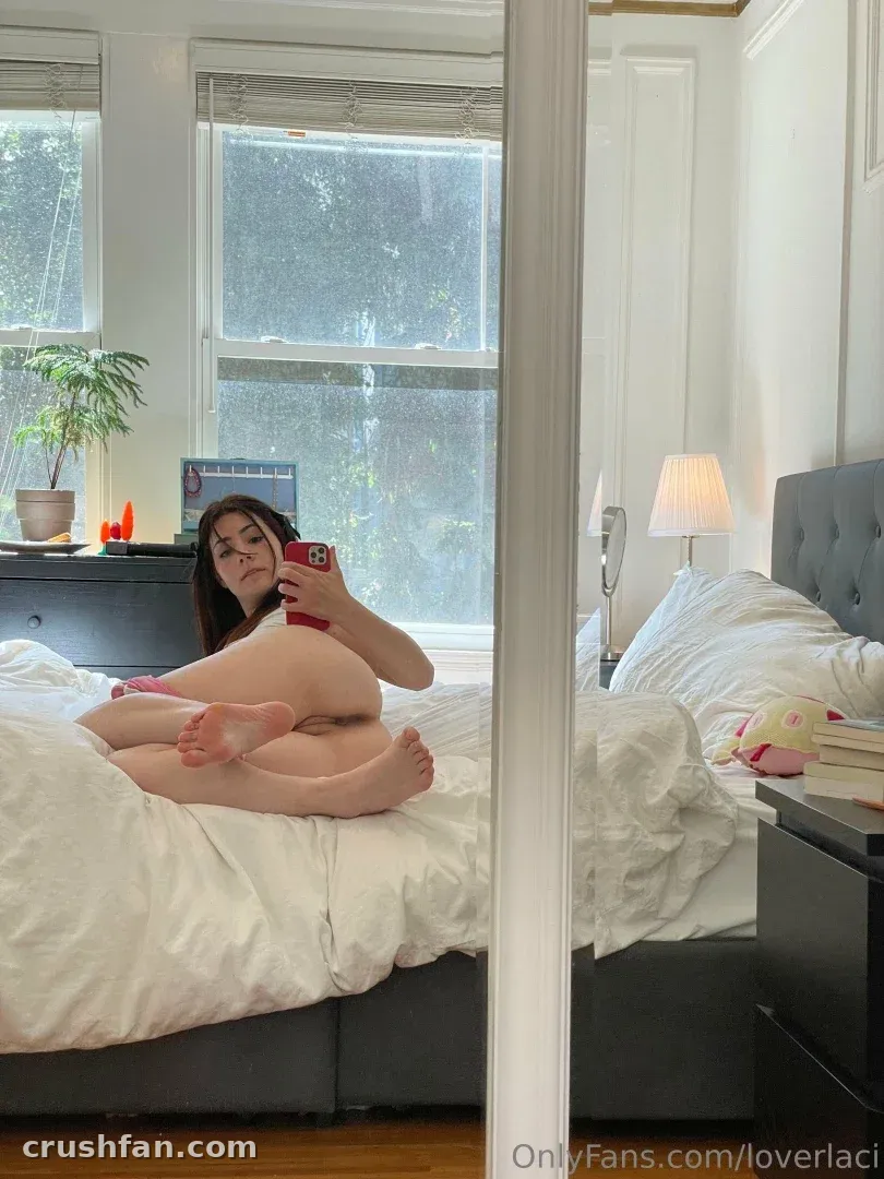 Laci Witton takes a mirror selfie while sitting on a white bed in a bright bedroom