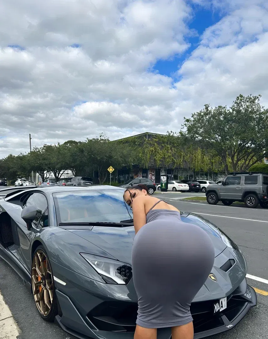 Woman in a grey dress leaning over a grey sports car in a sunny parking lot