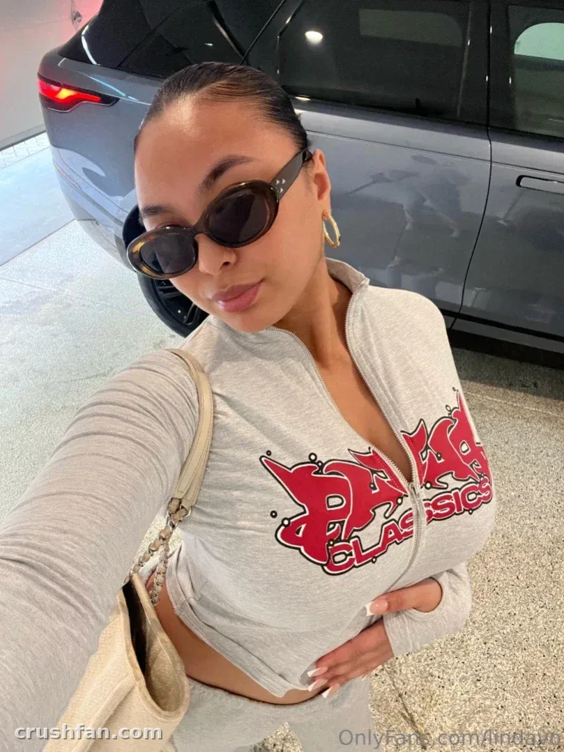 Linda Vo dans une veste grise à fermeture éclair et des lunettes de soleil prenant un selfie à côté d'une voiture sombre
