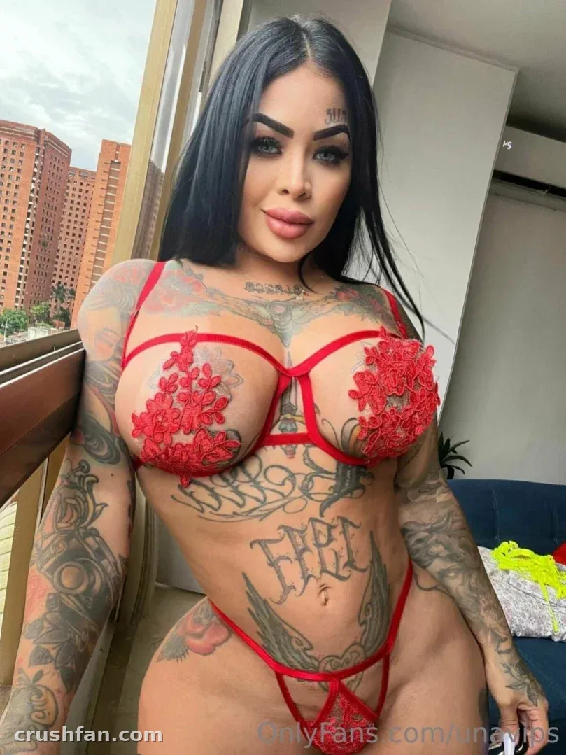 Una Cakez com lingerie de renda vermelha em pé junto a uma janela com vista da cidade