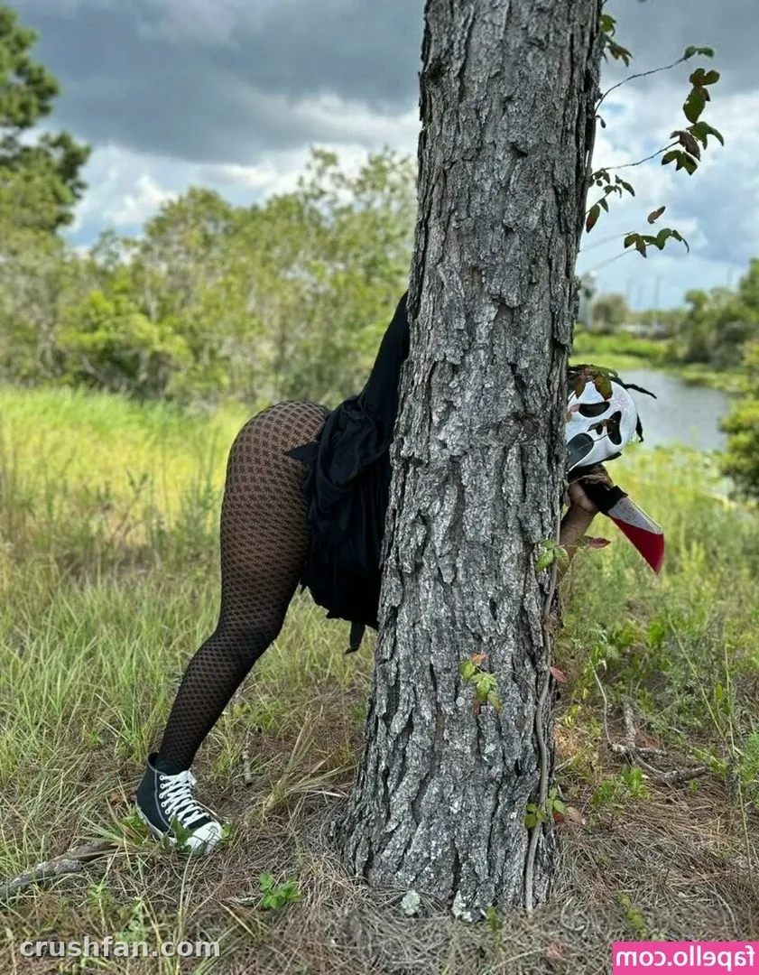 Creadora con falda negra, medias de rejilla y zapatillas de caña alta de pie detrás de un árbol en un campo de hierba