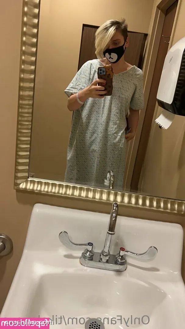 Tika com um vestido cinza texturizado tirando uma selfie no espelho acima de uma pia branca