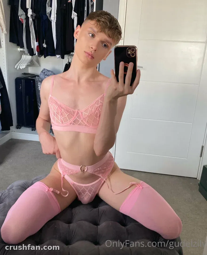 Criador com lingerie transparente rosa e meias tirando uma selfie no espelho enquanto está sentado em um otomano cinza.