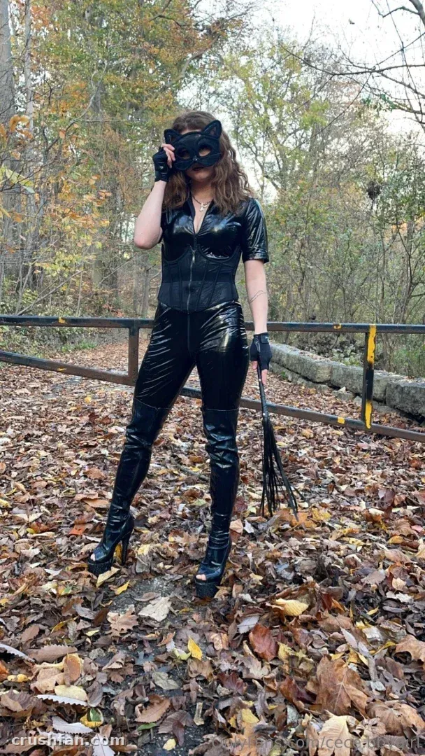 Creator in a black catsuit and cat mask standing on a leaf-strewn woodland path