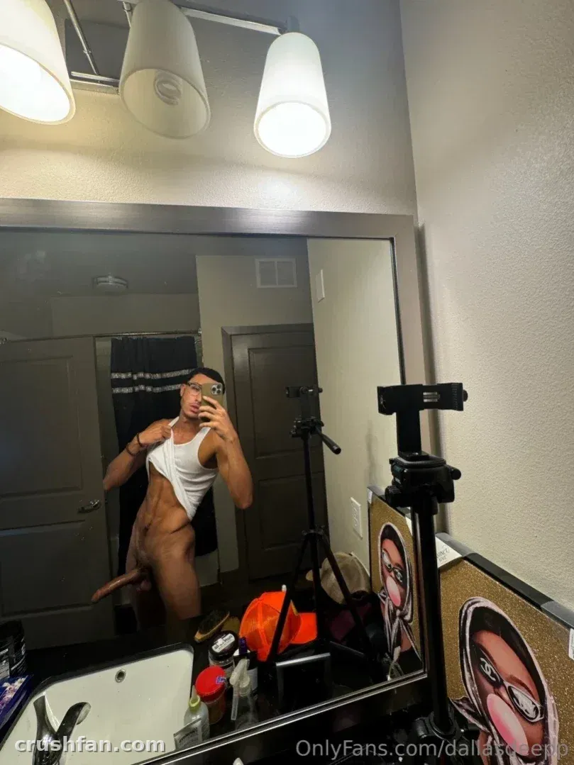 Dallas in a white tank top taking a mirror selfie in a bathroom with warm overhead lighting.