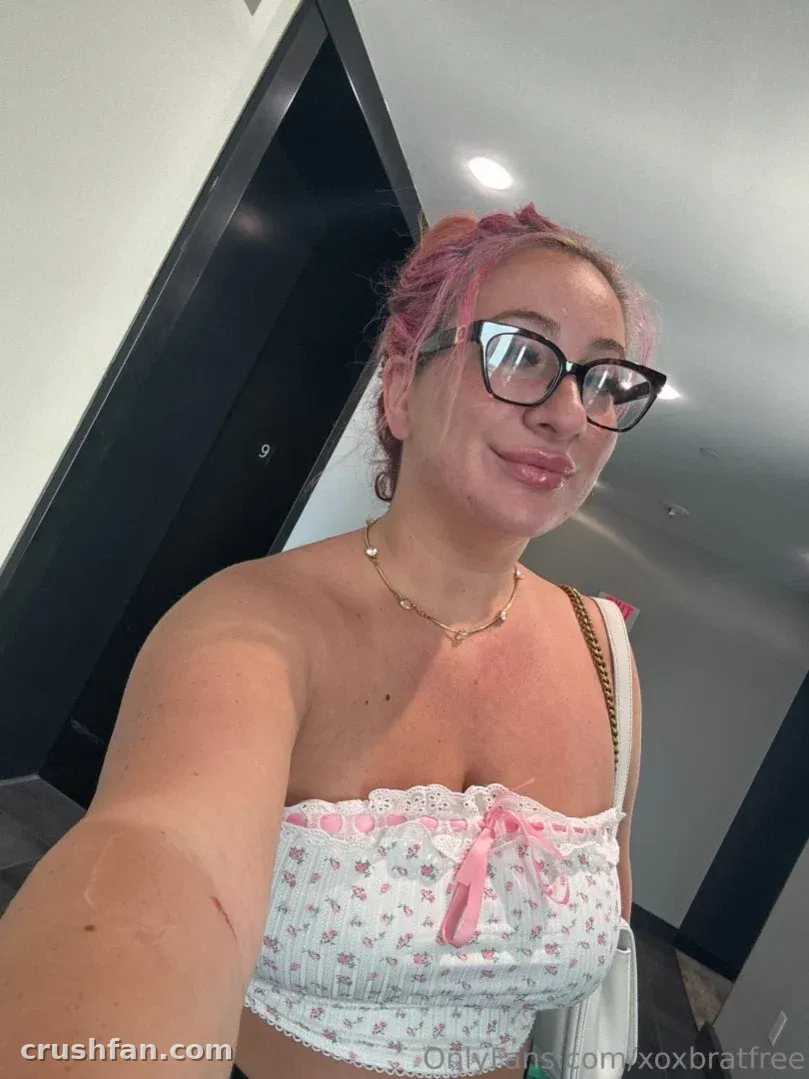 Criadora com cabelo rosa e óculos tira uma selfie clara em ambiente interno com um top floral.