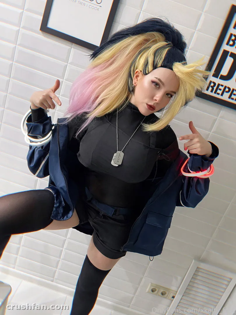 Creatorin in einem schwarzen Outfit und marineblauer Jacke posiert in einem gefliesten Raum mit pink- und blonden Haaren.
