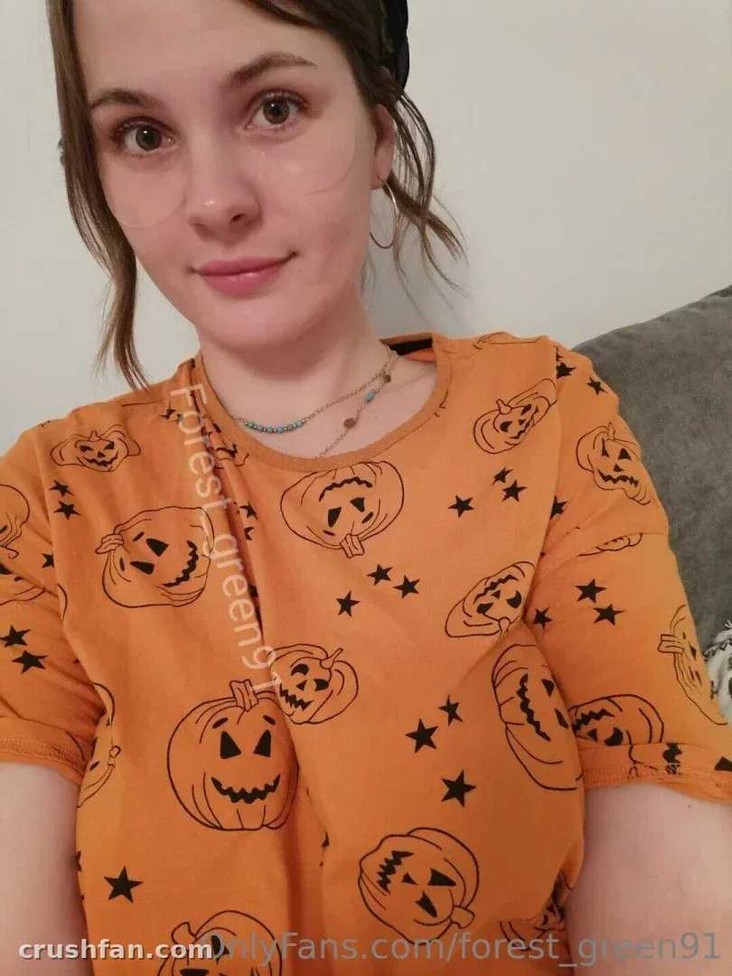Forest Green in einem orangefarbenen Top mit Kürbismuster posiert drinnen für ein weich beleuchtetes Selfie.