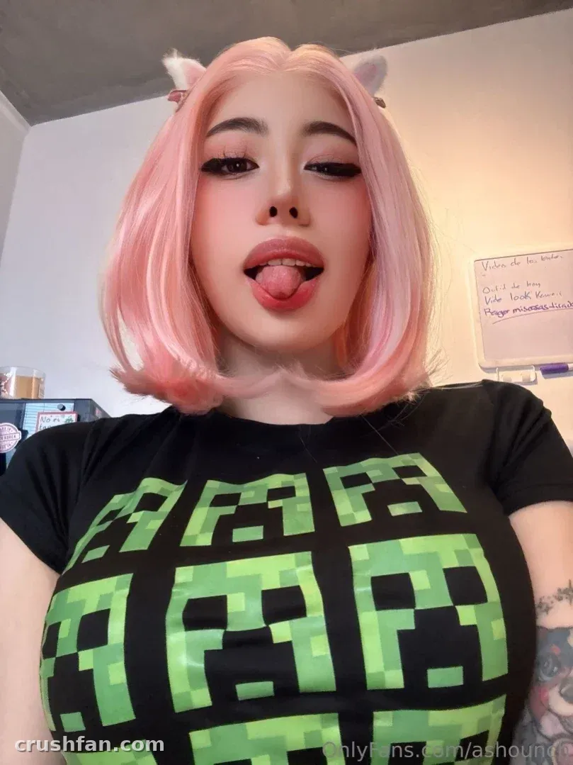 Asho con pelo rosa y orejas de gato sacando la lengua con un top de estampado de píxeles.