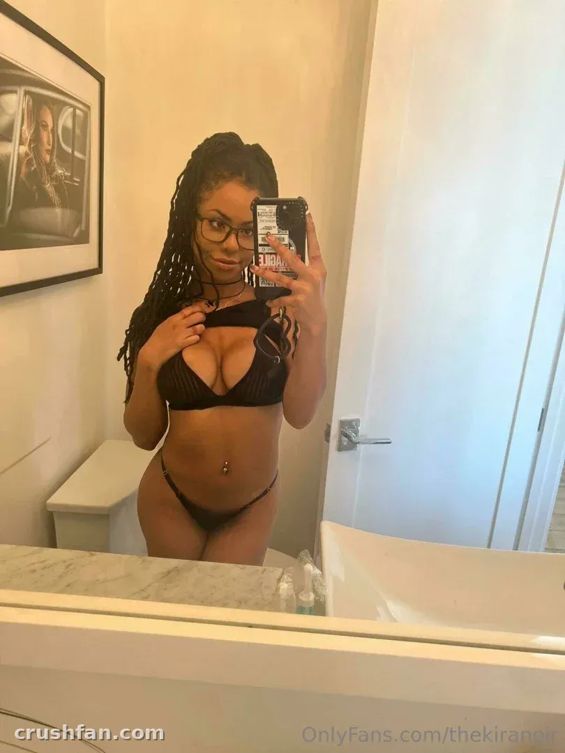 Kira Noir prenant un selfie dans le miroir dans une salle de bain lumineuse, portant un ensemble noir deux pièces.