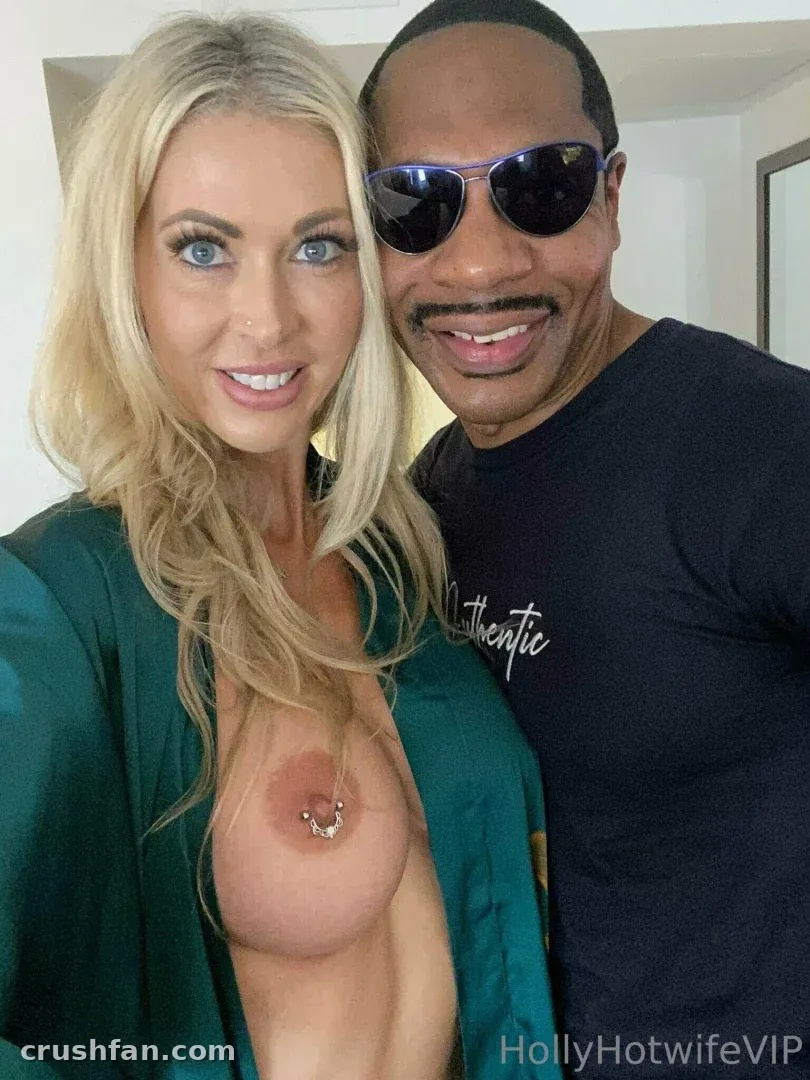 Holly Hotwife in einem grünen Morgenmantel neben einem Mann in einem schwarzen T-Shirt und Sonnenbrille in einem Innenraum