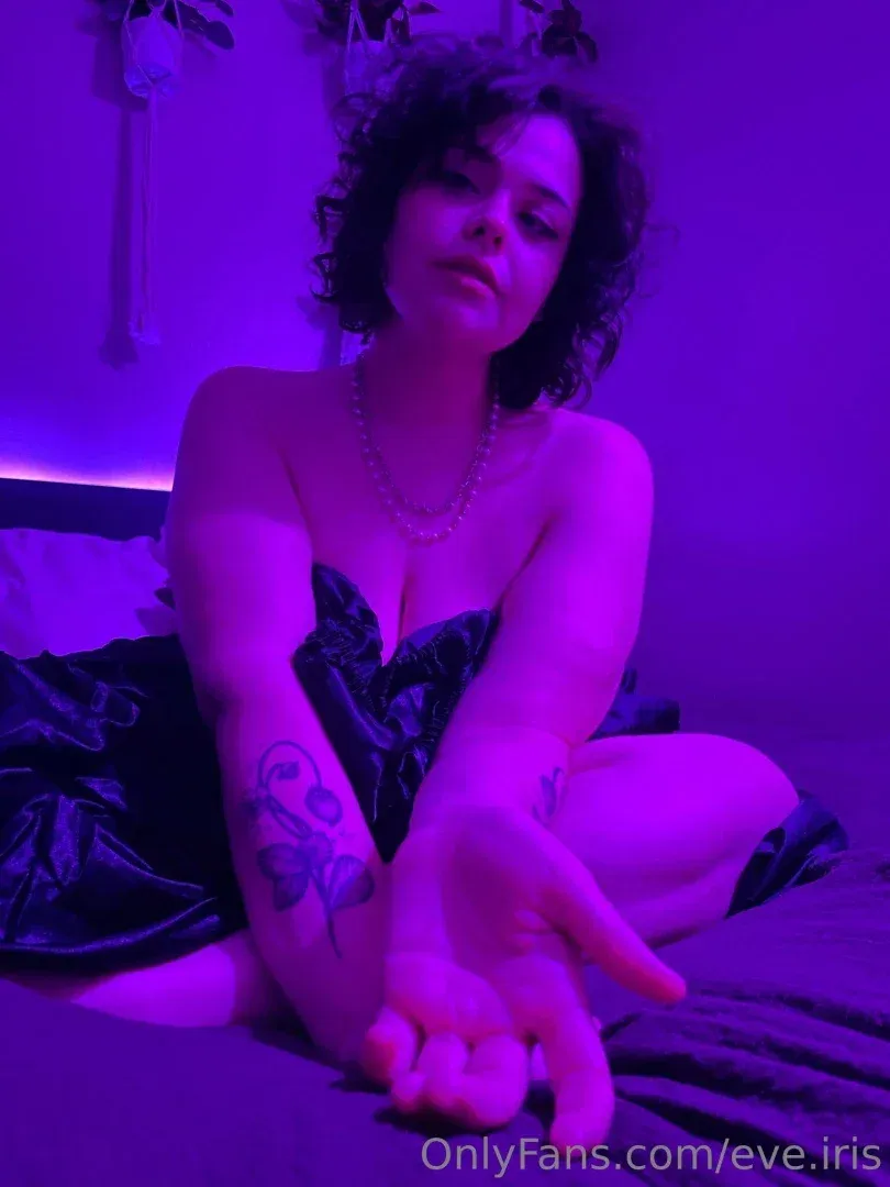 Eve Iris sentada en una cama bajo luz púrpura, con un vestido de satén oscuro y extendiendo la mano hacia la cámara.