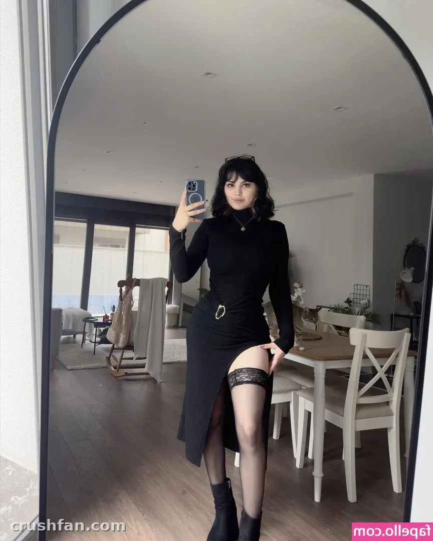 Ezra de vestido preto tira uma selfie no espelho em um ambiente claro com uma mesa de jantar.