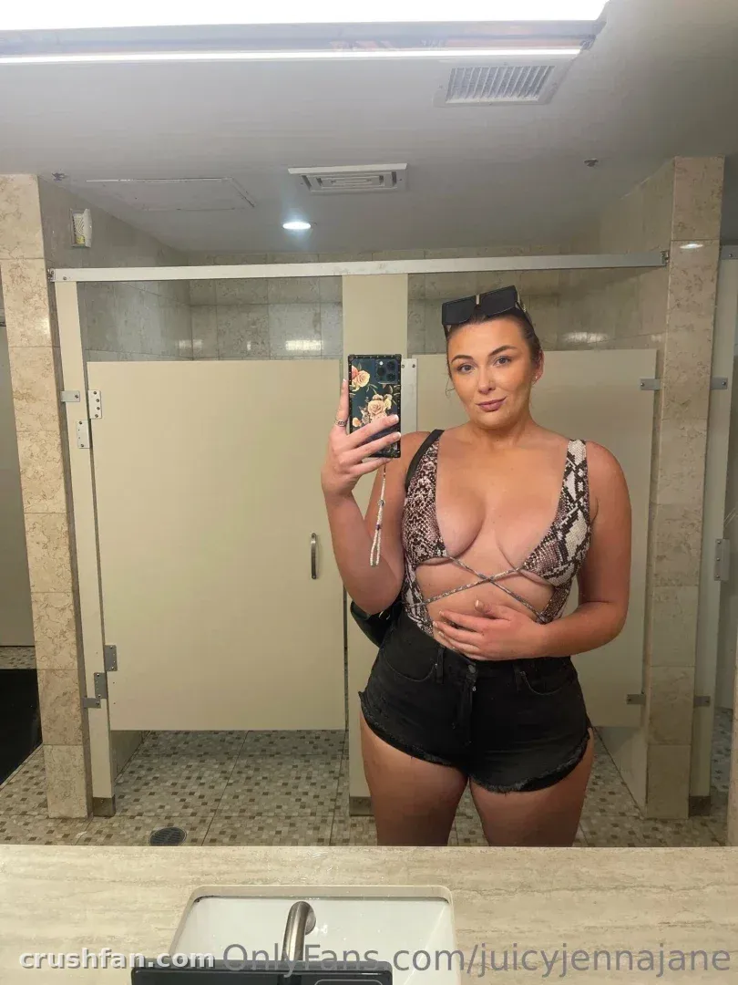 Jenna Jane en haut de bikini et short noir prenant un selfie dans le miroir dans des toilettes carrelées.