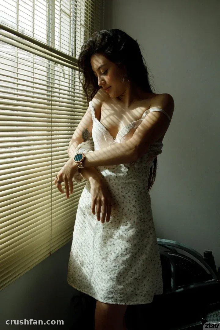 Ange in a floral dress standing by sunlit window blinds with striped shadows