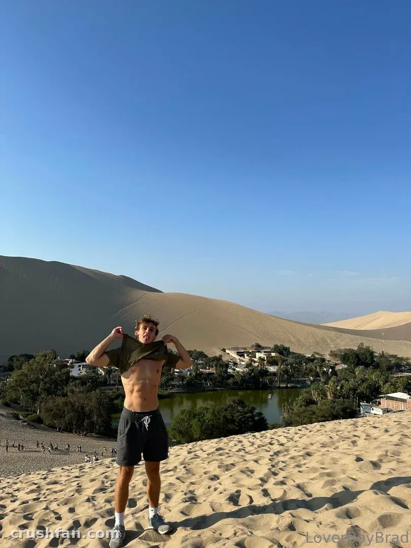 Leo levantándose la camiseta mientras está de pie en una duna de arena sobre una laguna bajo un cielo azul