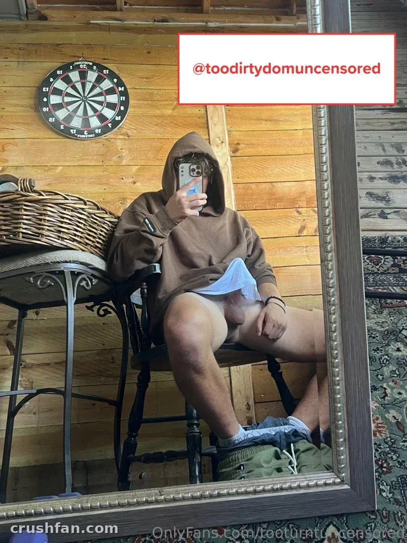 Too Turnt Tony com um moletom marrom tirando uma selfie no espelho em uma sala com painéis de madeira e um alvo de dardos.